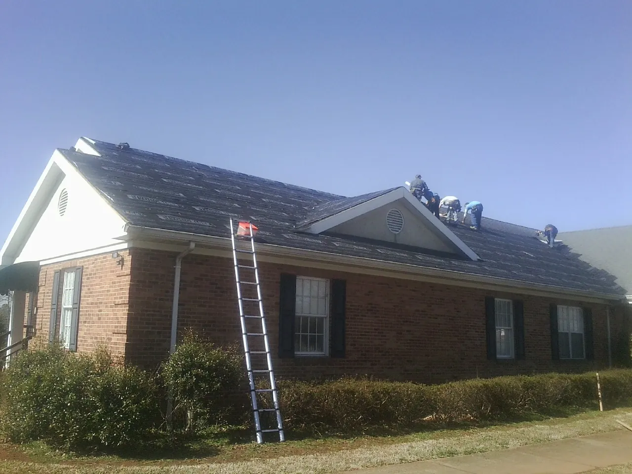 Crew tearing shingles off a brick ranch roof with a ladder set — Emergency Roof Repair work by our crew in Ether