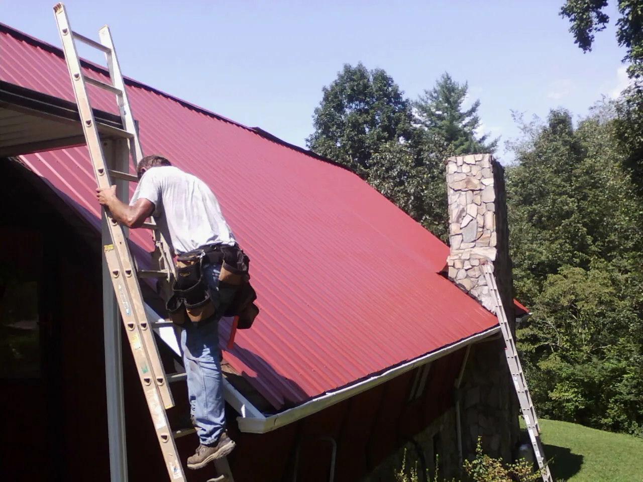 Roofer on a ladder beside a red metal roof and stone chimney — Soffit & Fascia Repair by Roofing Brothers in Ether